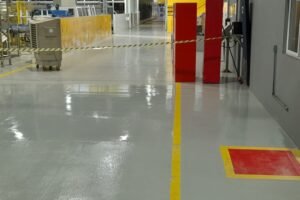 Fotografia do interior de uma fábrica destacando um Piso Epóxi Industrial cinza de alto brilho, com linhas amarelas e marcações vermelhas no chão para sinalização de rotas logísticas e segurança.