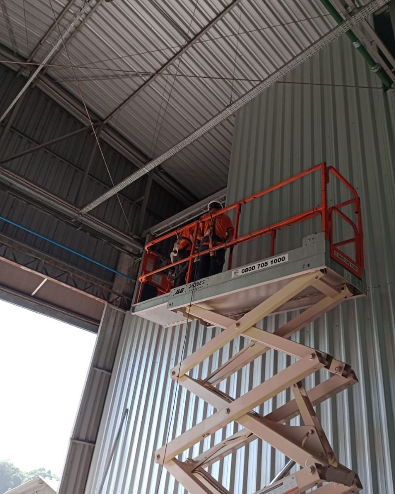 Equipe da Emkorp utilizando plataforma elevatória tipo tesoura para instalação de infraestrutura técnica e eletrocalhas no teto de um galpão industrial em Joinville.