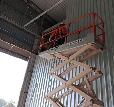 Equipe de engenharia da Emkorp utilizando plataforma elevatória tipo tesoura para realizar instalação de infraestrutura técnica em altura dentro de um galpão industrial em Joinville.