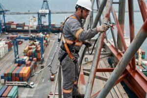 Trabalhador com EPIs e cinto de segurança realizando pintura anticorrosiva no alto de um guindaste de terminal portuário, com pátio de contêineres ao fundo.