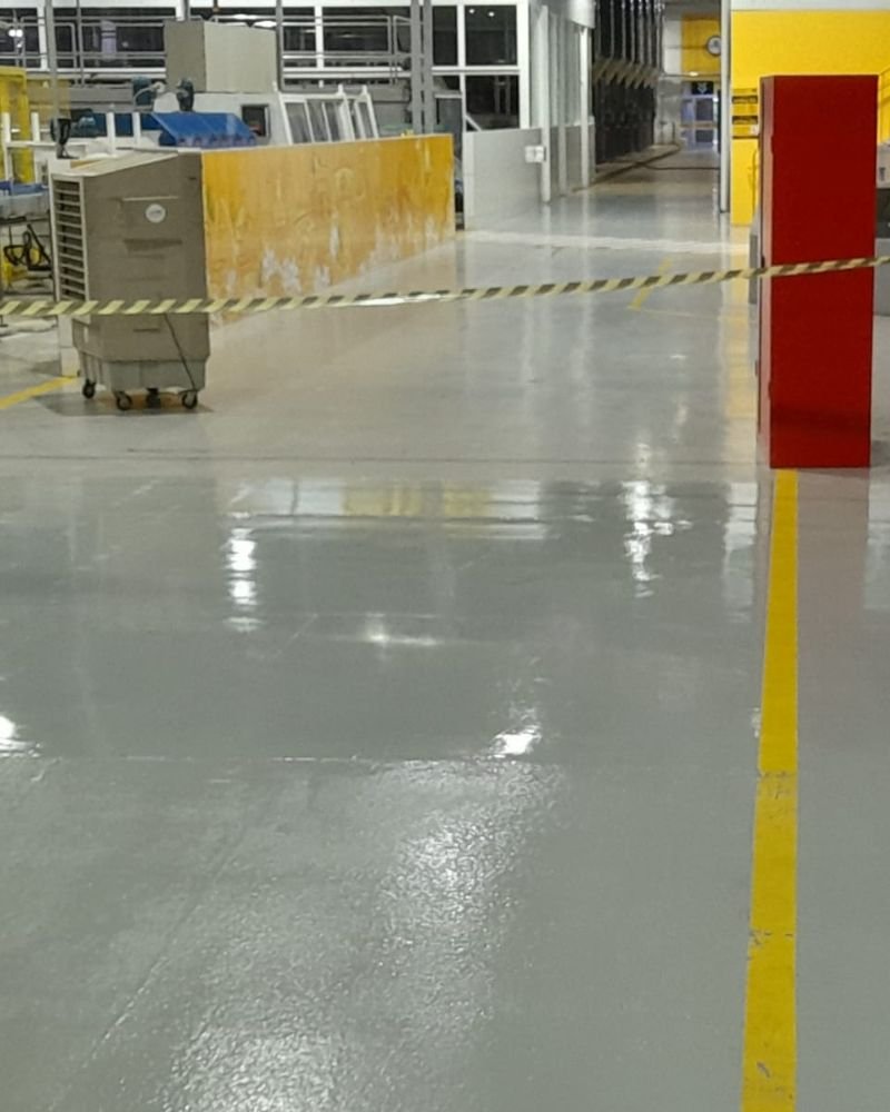 Piso industrial com revestimento epóxi cinza brilhante e faixa de demarcação de segurança amarela, recém-aplicado pela engenharia da Emkorp em Joinville, com área isolada para cura rápida.