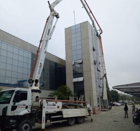 Caminhão bomba de concreto operando com braço articulado em um edifício comercial moderno com fachada de vidro, durante obra de reforma corporativa e retrofit executada pela Emkorp em Joinville.
