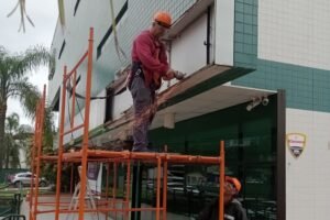 Dois trabalhadores em um andaime realizando obras de Retrofit Corporativo na fachada de um prédio comercial com revestimento de pastilhas verdes.