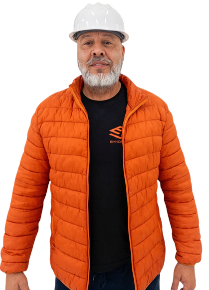 Retrato de Sandro Kindermann, diretor e responsável técnico da Emkorp Empreendimentos, vestindo capacete branco de engenharia, jaqueta laranja e camiseta preta com a logo da empresa, em fundo escuro.